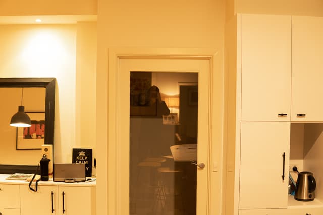 A warmly lit kitchen interior with white cabinets, a kettle, and a view into an adjoining room