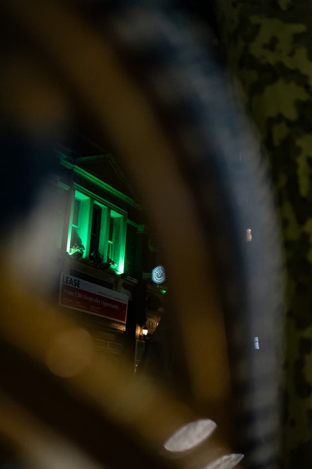 A nighttime scene viewed through a circular frame, possibly a wheel, with a focus on a green-lit window and a blurred street sign in the background