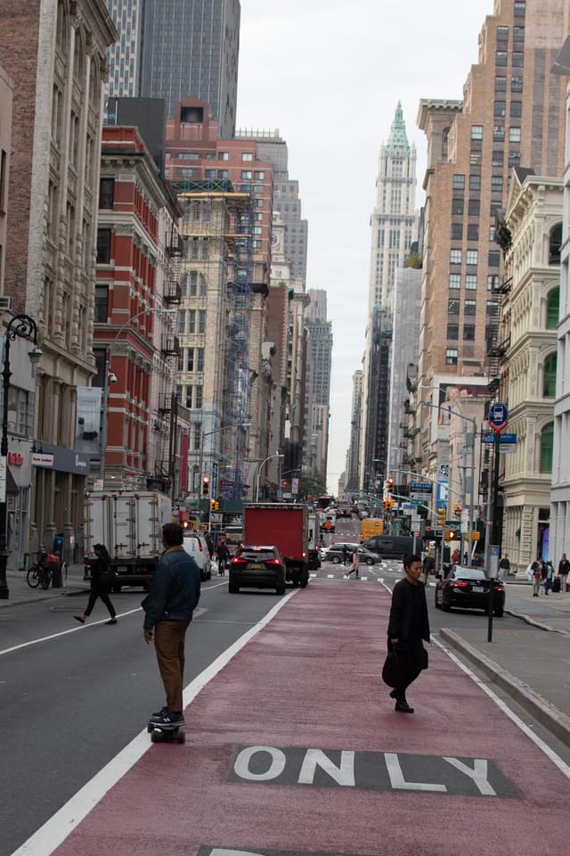 A bustling city street with a person skateboarding and another walking, tall buildings lining the road, and a red bus lane marked ONLY