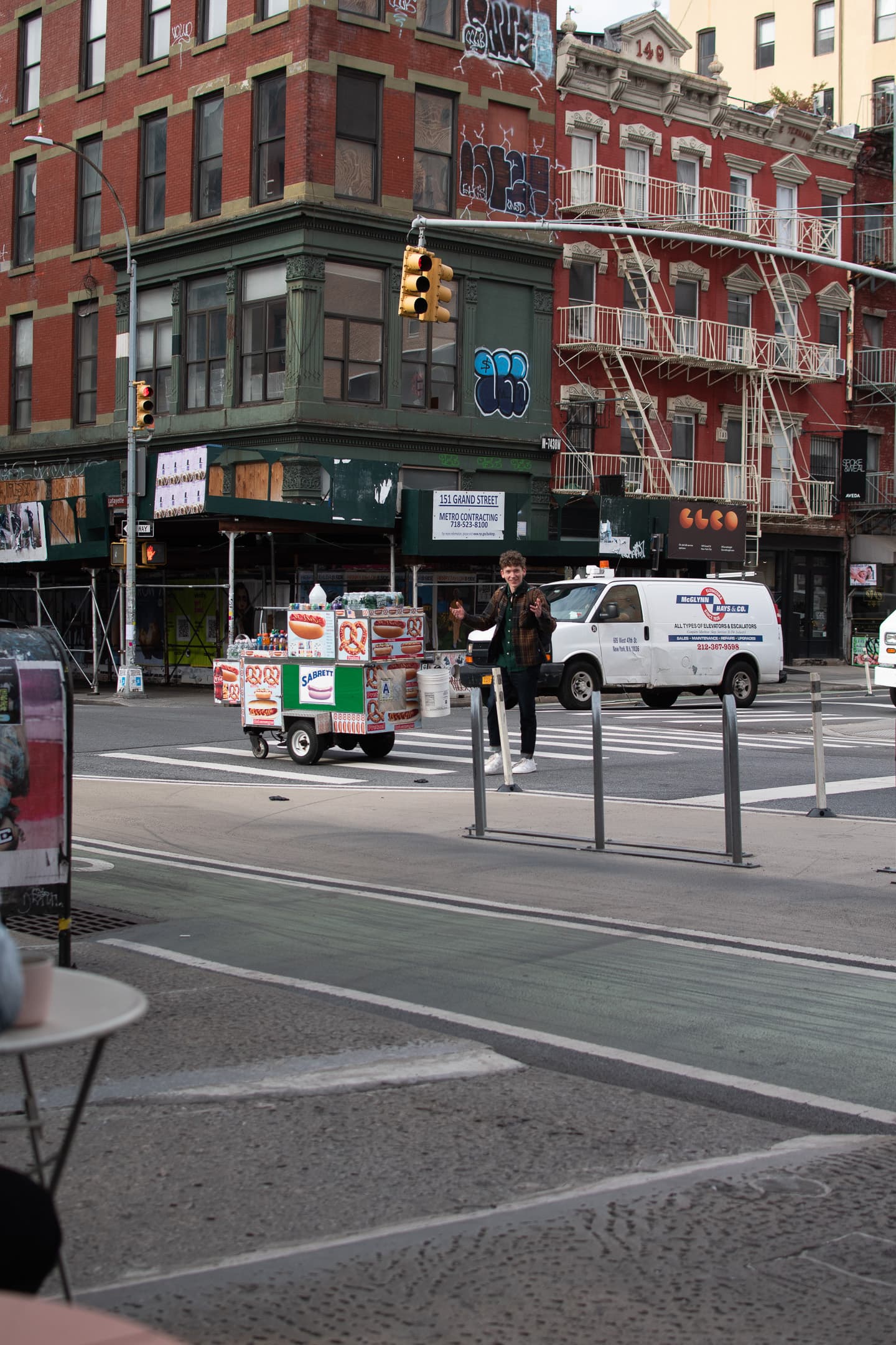 A bustling city street corner with a pedestrian crossing, a food cart, vehicles, and colorful buildings with storefronts