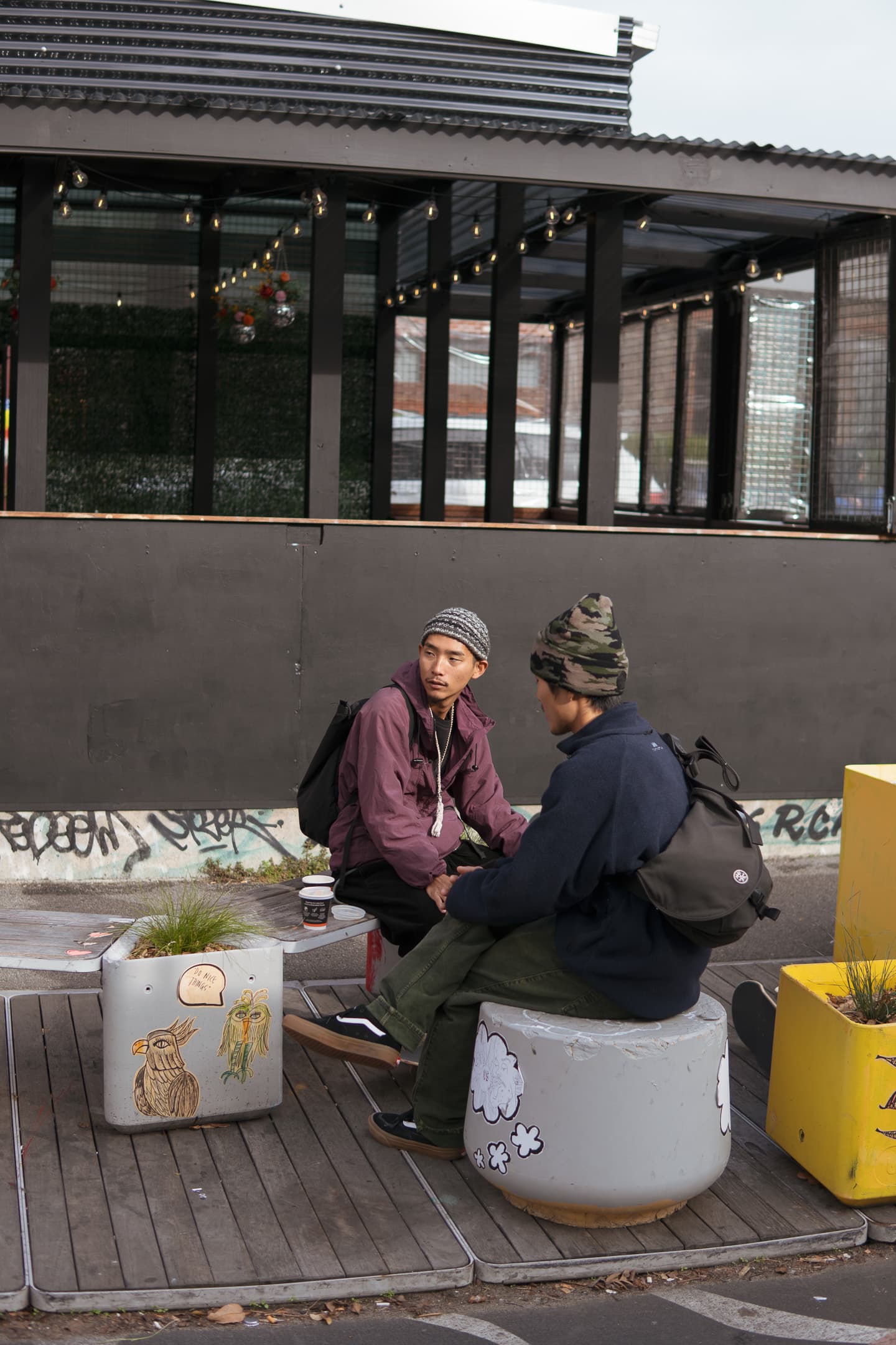 Two individuals are seated on makeshift stools outdoors, engaged in conversation. The setting appears to be urban with graffiti on a wall in the background and a modern building structure behind them. The stools they are sitting on are creatively decorated with various designs