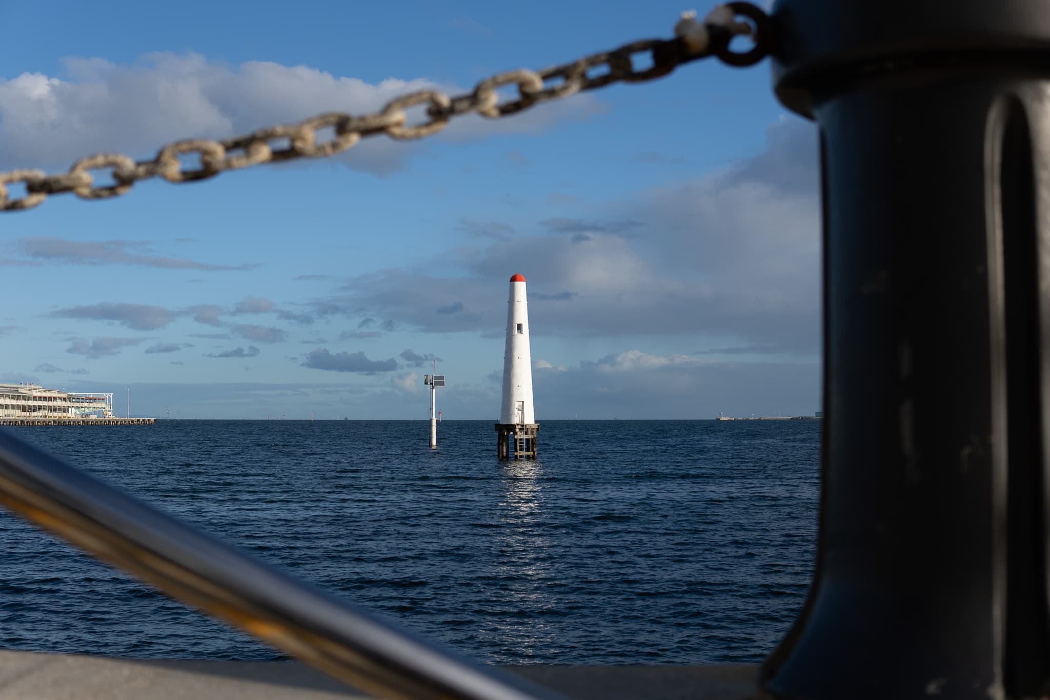 A serene seascape framed by a metal chain and post, featuring a distant sailboat and a clear blue sky with scattered clouds