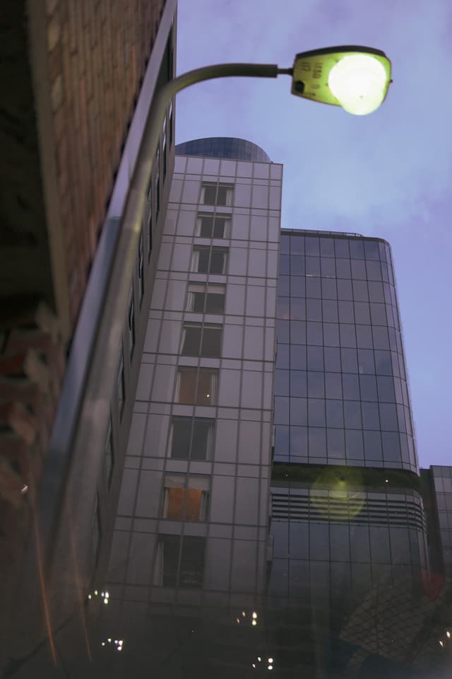 A lit street lamp in the foreground with a modern glass building in the background, captured during dusk or dawn