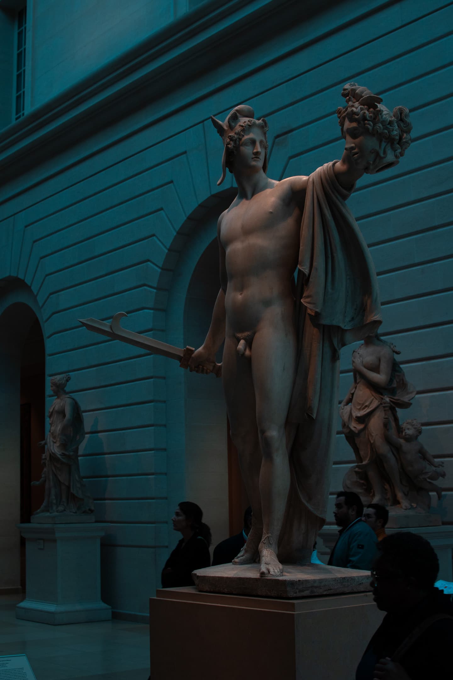 A classical statue, likely representing a mythological figure, stands prominently in a dimly lit museum gallery with visitors observing the artwork