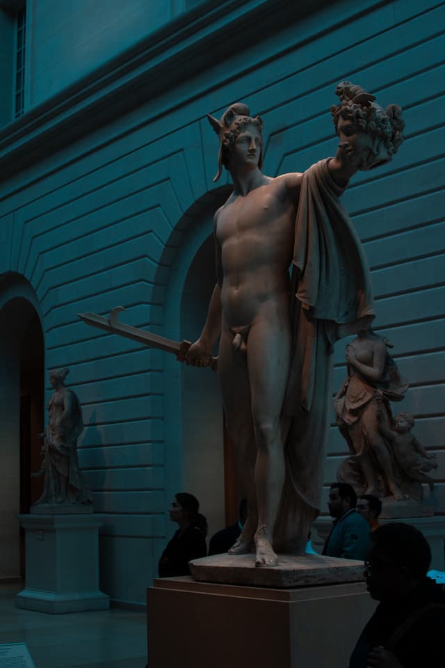 A classical statue, likely representing a mythological figure, stands prominently in a dimly lit museum gallery with visitors observing the artwork