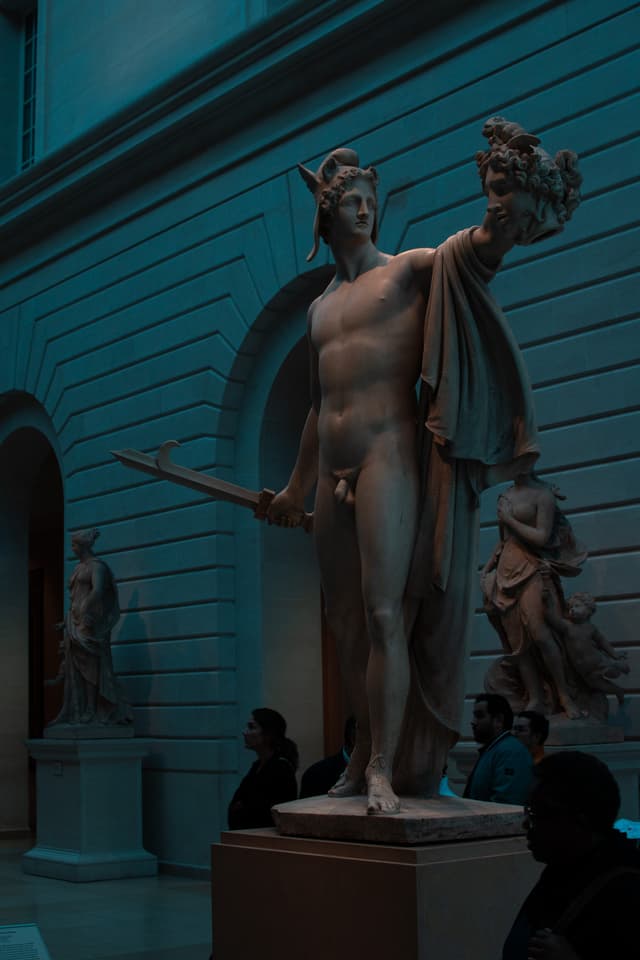 A classical statue, likely representing a mythological figure, stands prominently in a dimly lit museum gallery with visitors observing the artwork