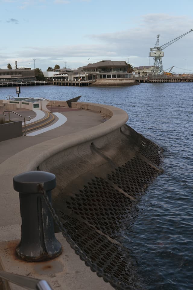 A curved waterfront promenade with a metal bollard in the foreground, overlooking a calm harbor with a crane in the distance