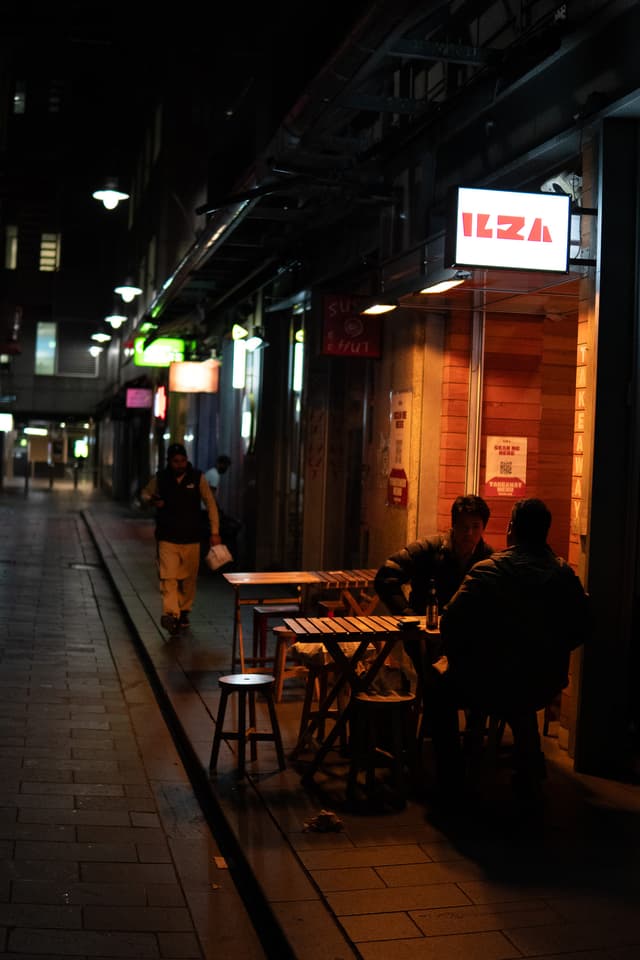 A dimly lit street at night with a person walking and a small group sitting at a table outside a shop with a lit sign