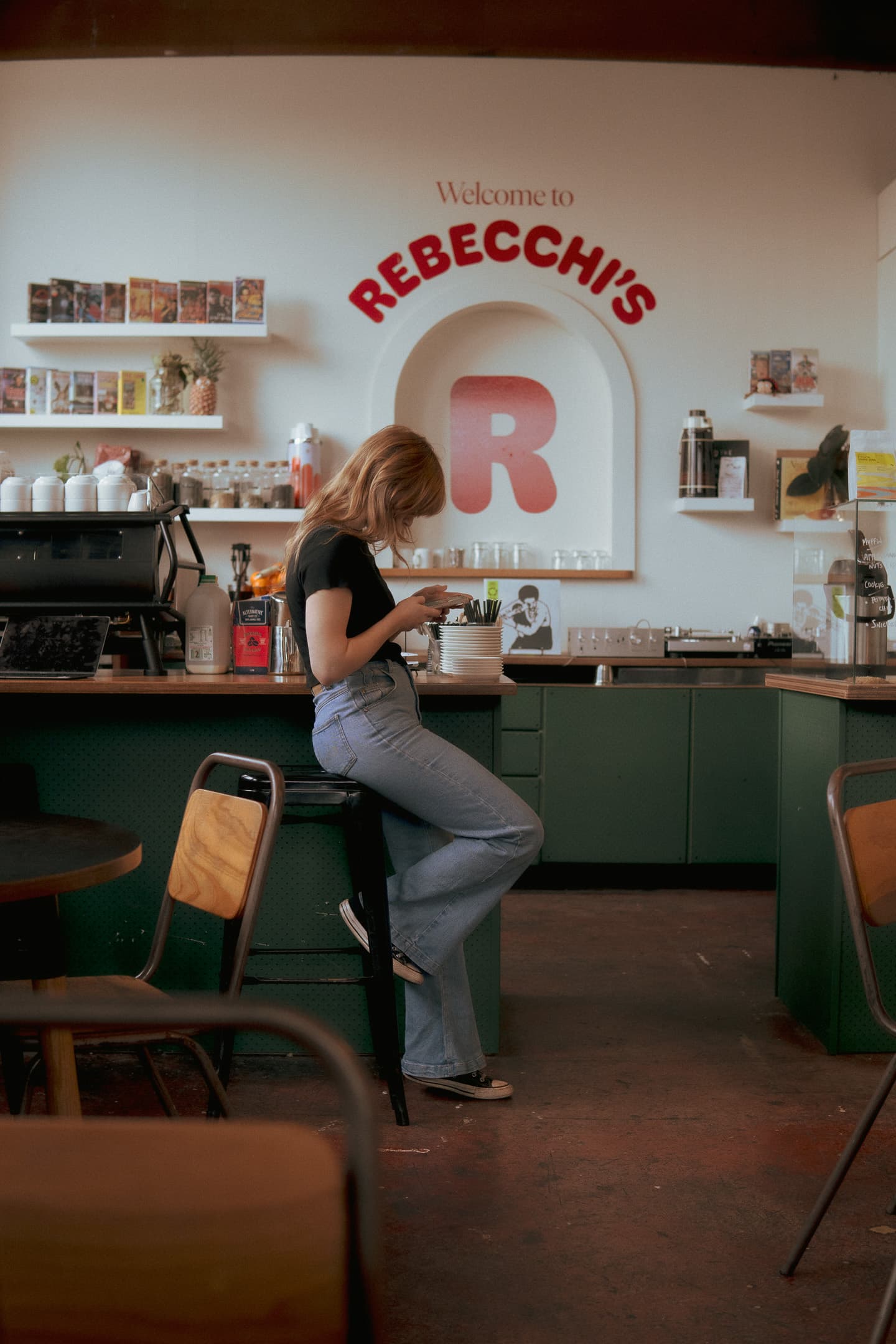 A woman is standing at a counter in a cafe named REBECCHI'S, holding a cup, possibly engaged in making a drink The cafe has a vintage aesthetic with a prominent red R on the wall