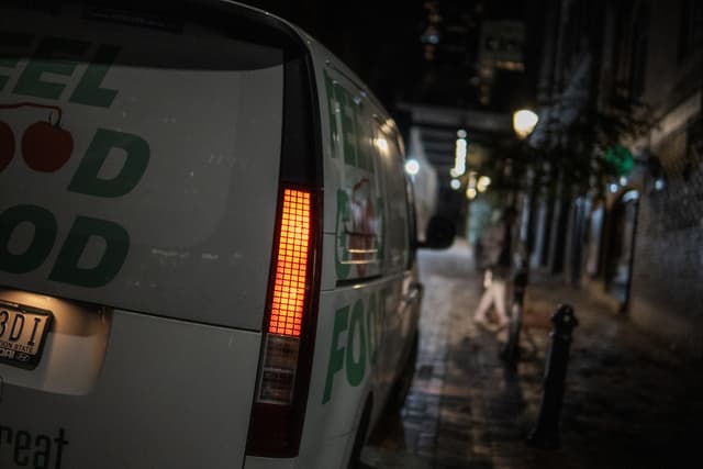 A van with the words FOOD written on it is parked on a wet street at night, with street lamps illuminating the scene and a person walking in the background