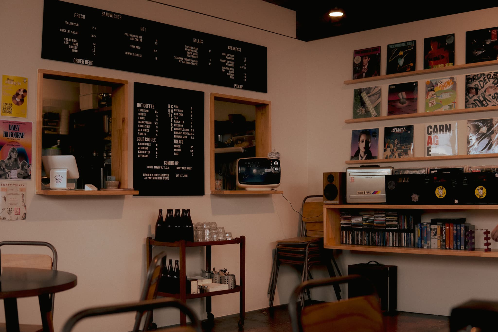 A cozy café interior with a menu board, shelves of books and records, a seating area with chairs, and a warm, inviting ambiance