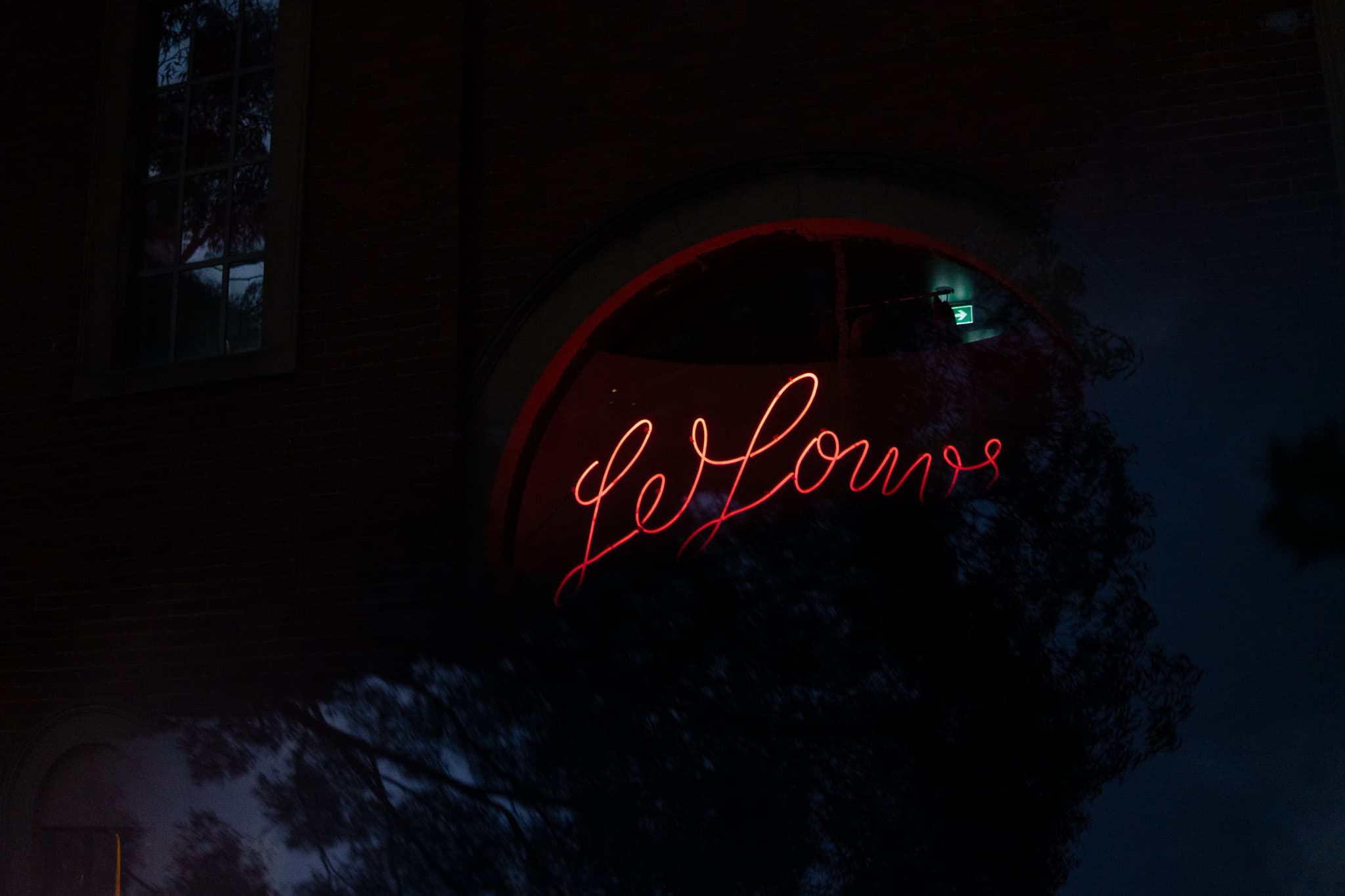 A neon sign with cursive lettering is mounted on a dark building facade, glowing in a reddish hue against the evening sky