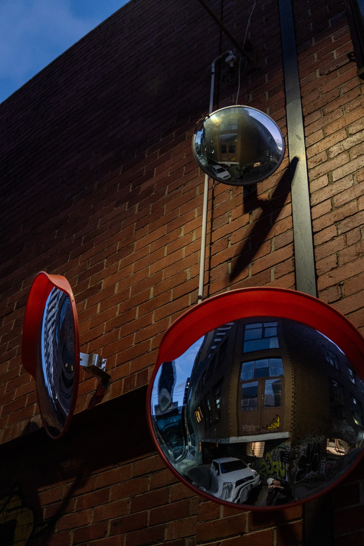 A pair of convex mirrors mounted on a brick wall, reflecting the urban environment around them, with a clear sky fading into dusk in the background