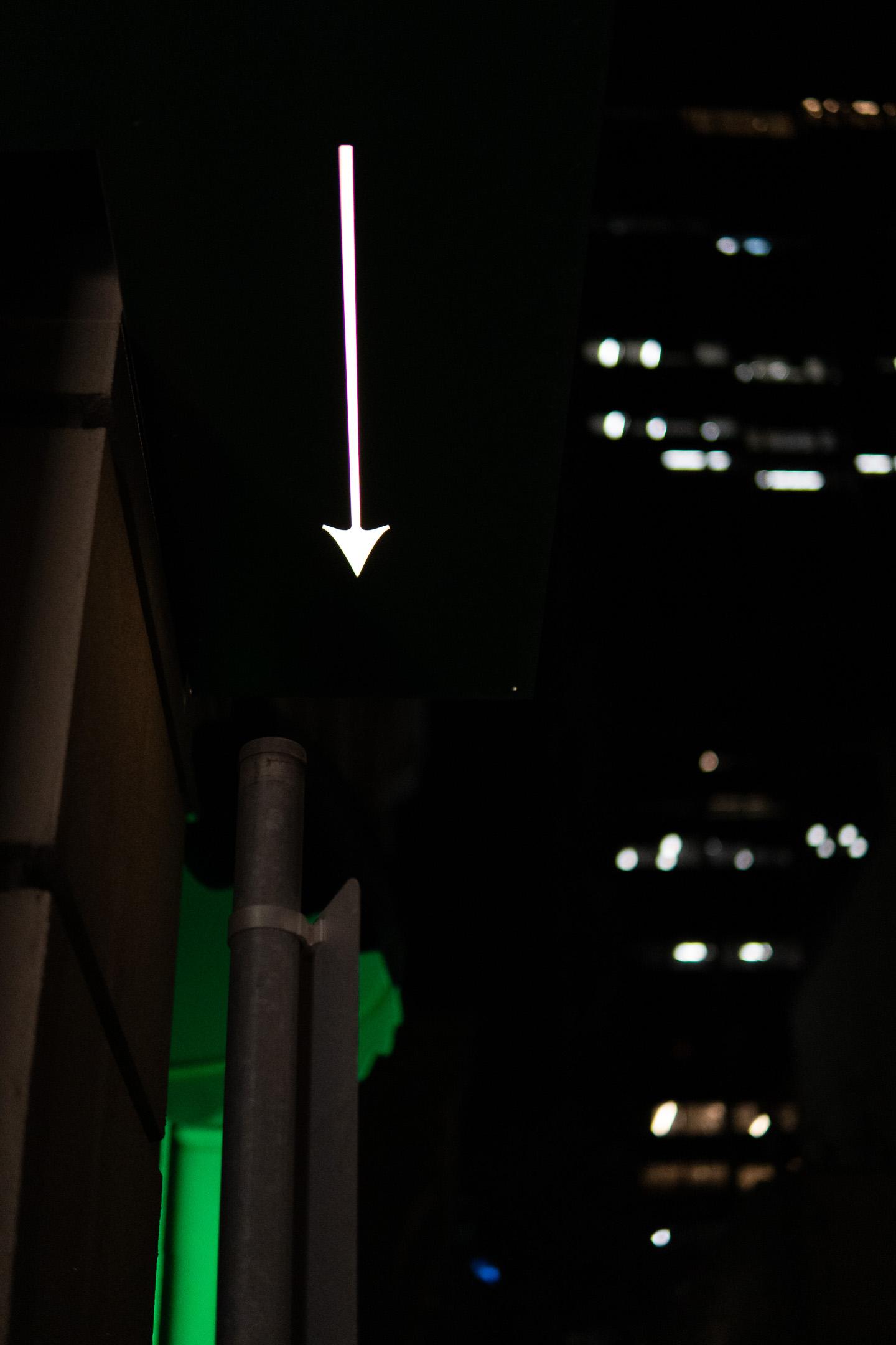 A neon arrow pointing downward against a dark urban backdrop with blurred building lights in the distance