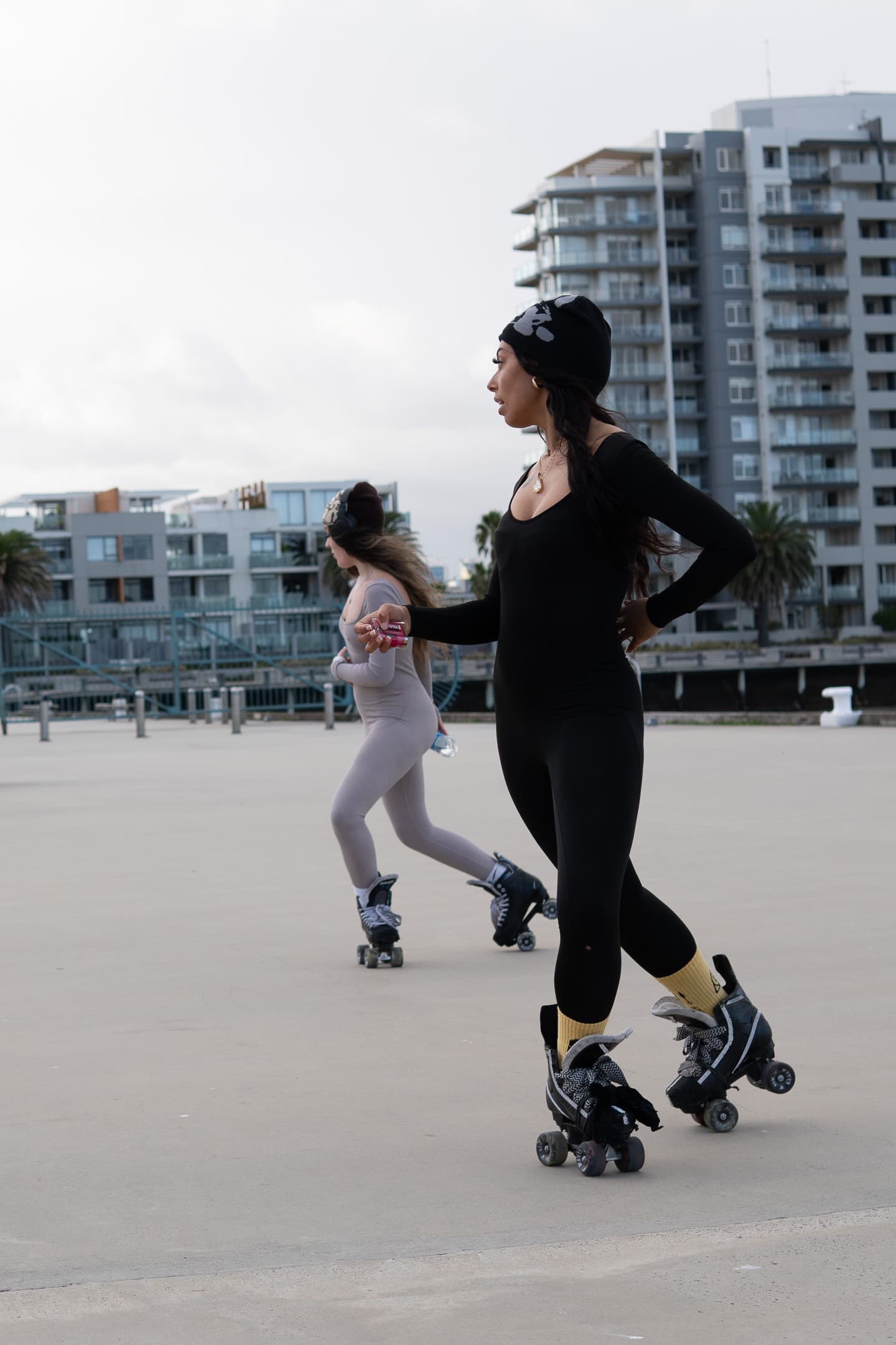 Two people are rollerblading on a smooth outdoor surface with buildings in the background. The person in the foreground is wearing black attire and rollerblades with yellow wheels, while the person in the background is dressed in white and pink, with white rollerblades