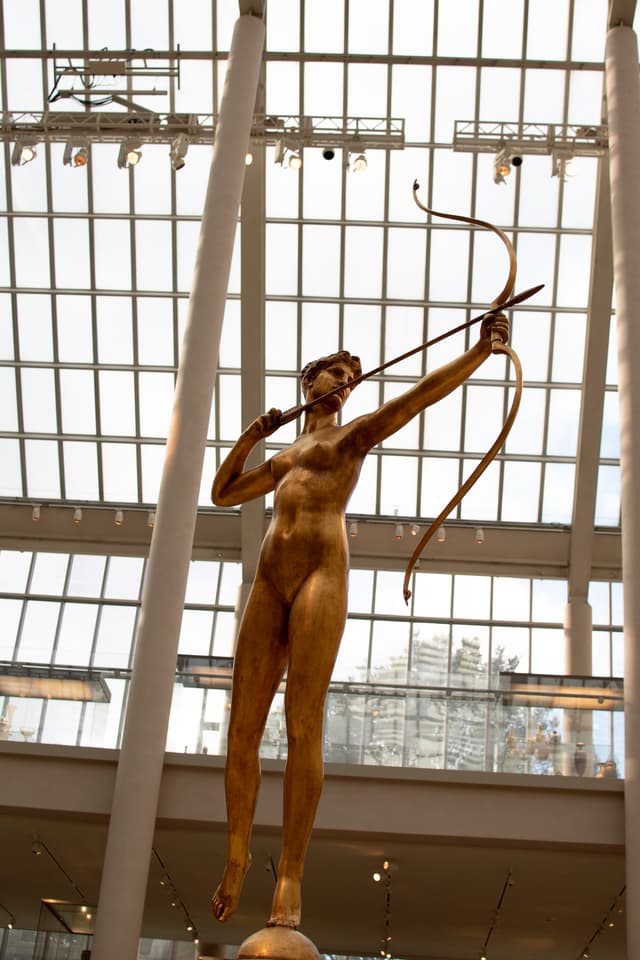 A statue of a female figure with a bow, poised as if ready to shoot an arrow, displayed in a gallery with a glass ceiling and structural beams