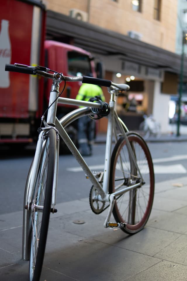 A bicycle is parked on a city sidewalk with a blurred background featuring a person in a high-visibility jacket and a red truck