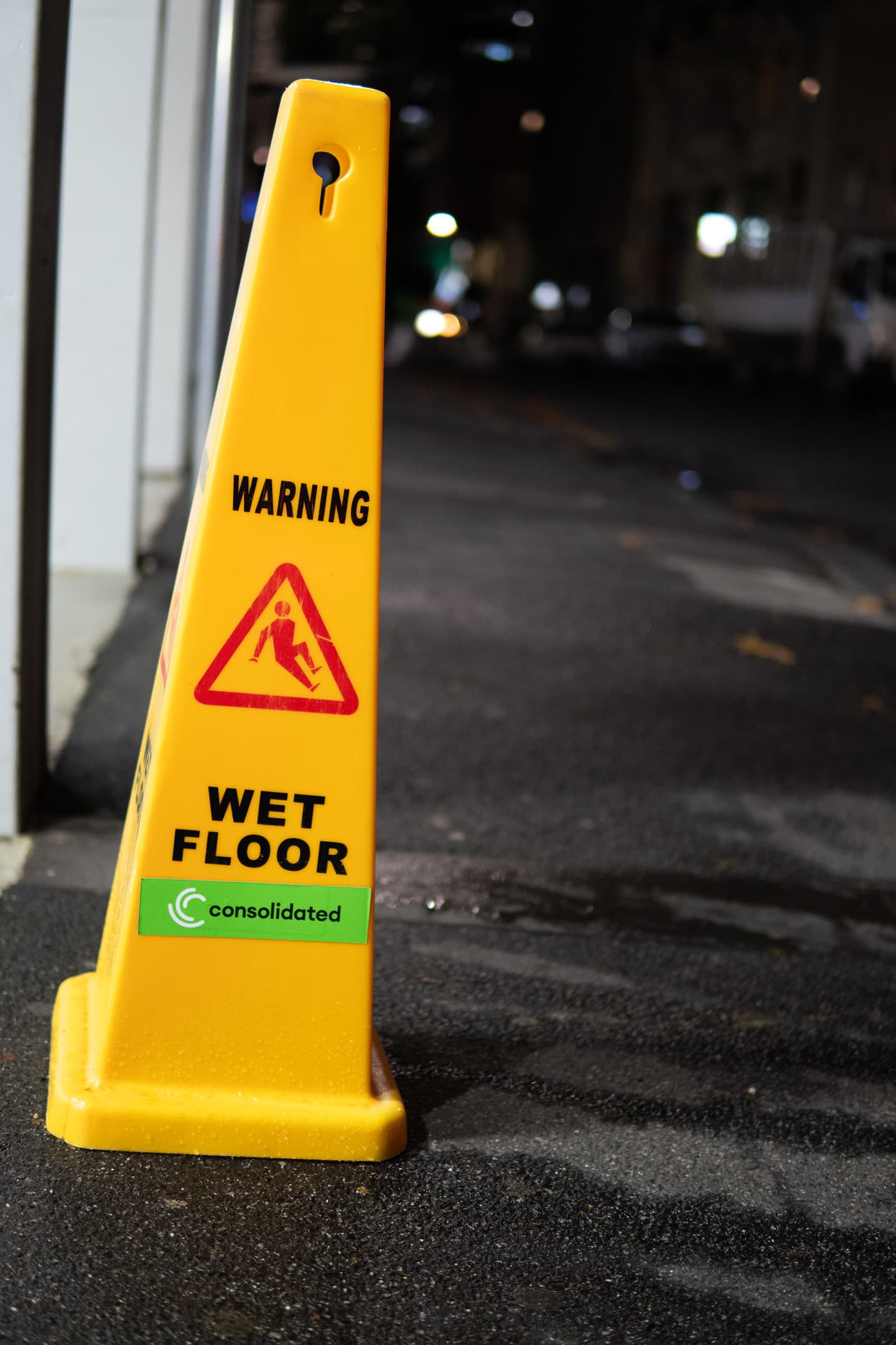 A yellow caution sign on a sidewalk at night, warning of a wet floor with a red triangle symbol indicating caution