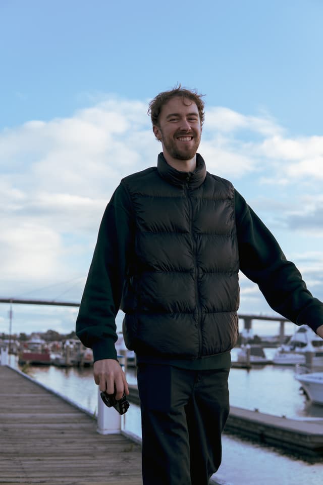 A smiling man in a black vest and trousers stands on a wooden dock with boats and water in the background under a partly cloudy sky. He is holding a bottle in his right hand