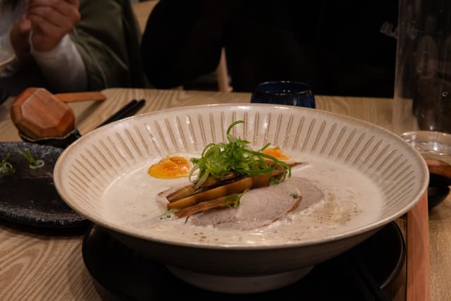 A plated dish is presented on a table, featuring what appears to be a gourmet preparation with a garnish on top. In the background, there are blurred elements including a person's hands and drinking glasses, suggesting a dining environment