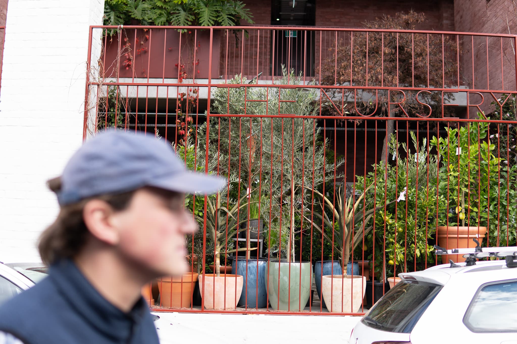 A person in a cap is in the foreground, slightly out of focus, with a balcony and potted plants in the background