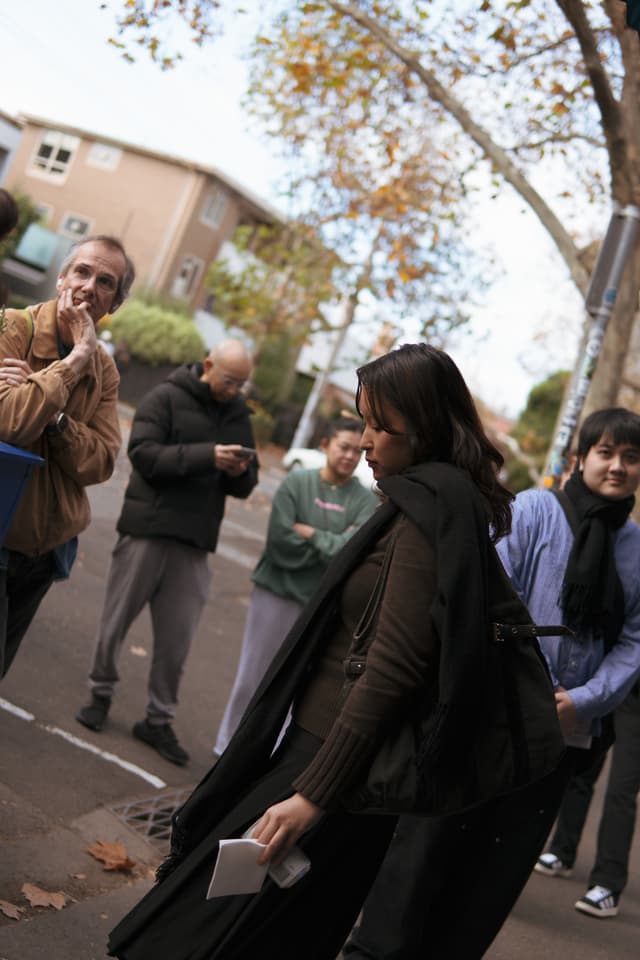 A group of people standing outdoors, with a woman in the foreground walking past and a man in the background appearing to be speaking and gesturing with his hand near his mouth