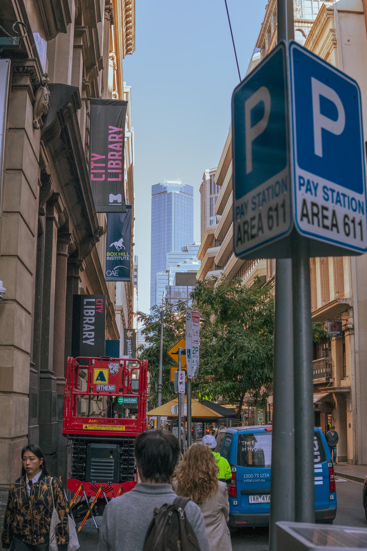 A bustling city street scene with pedestrians, a parking sign, and a cherry picker, with skyscrapers visible in the background under a clear sky