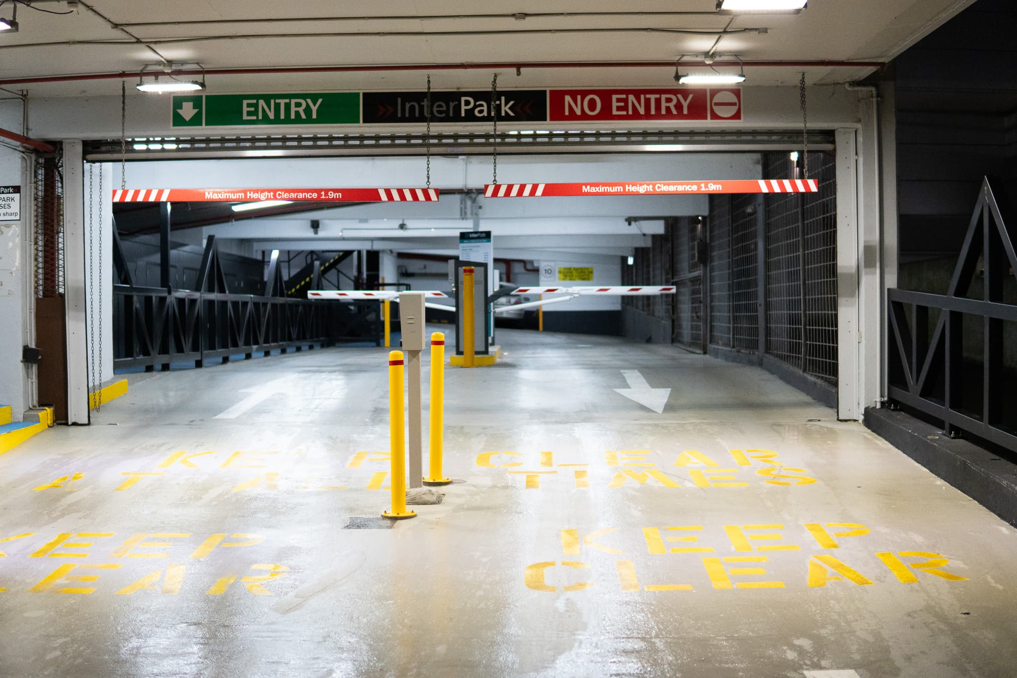 An empty parking garage entrance with a green ENTRY sign above an open gate and a red NO ENTRY sign above a closed gate, directional arrows on the ground, and a yellow safety column in the foreground