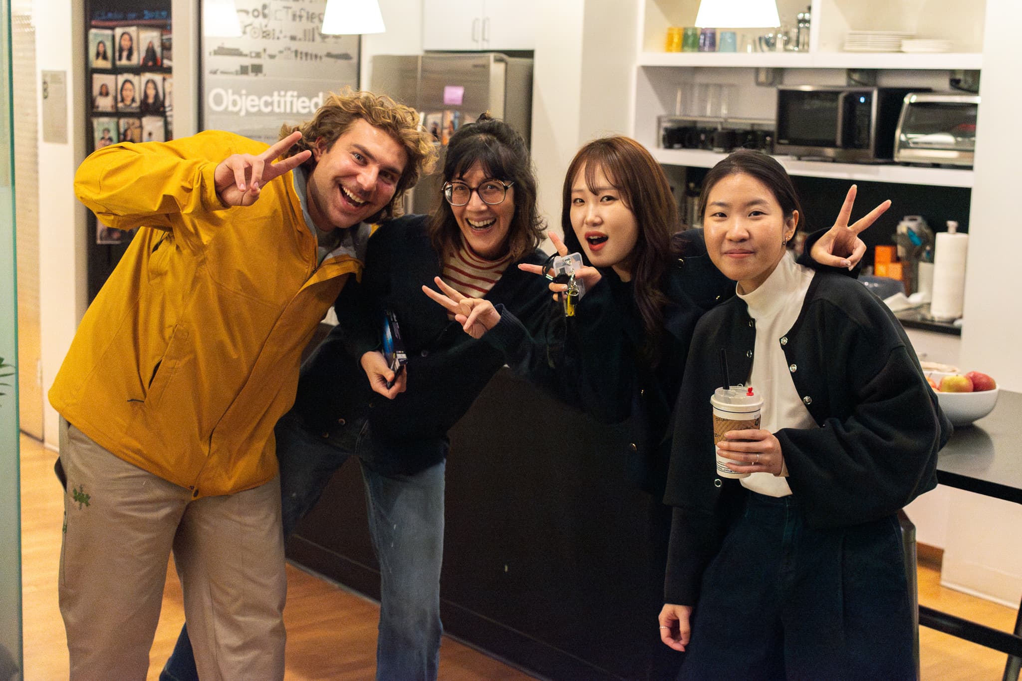 Four people are posing for a photo in a kitchen, smiling and making peace sign gestures