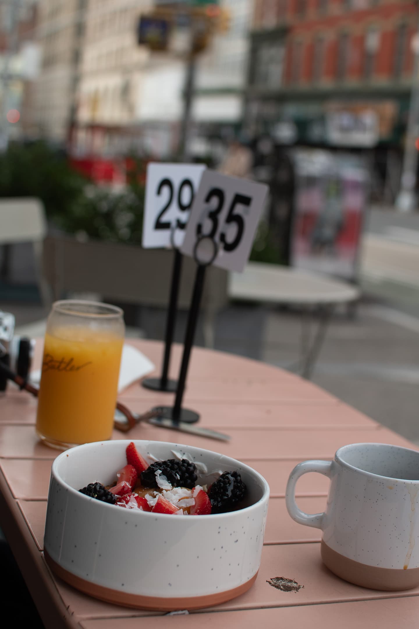 A breakfast setup on an outdoor table featuring a bowl of yogurt with berries, a glass of orange juice, and a coffee cup, with table number signs in the background