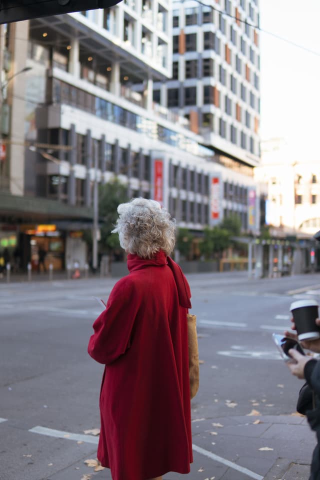 A person with curly hair wearing a red coat stands on a city street, facing away from the camera