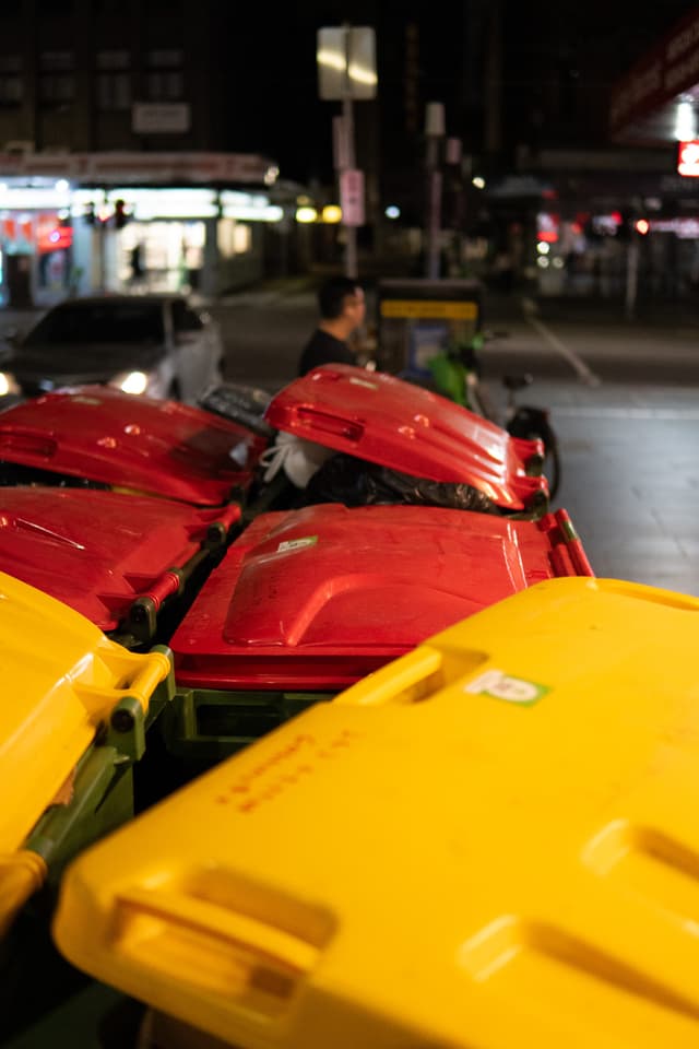 A nighttime street scene with a row of brightly colored red and yellow waste bins in the foreground and a person standing in the background amidst urban lighting