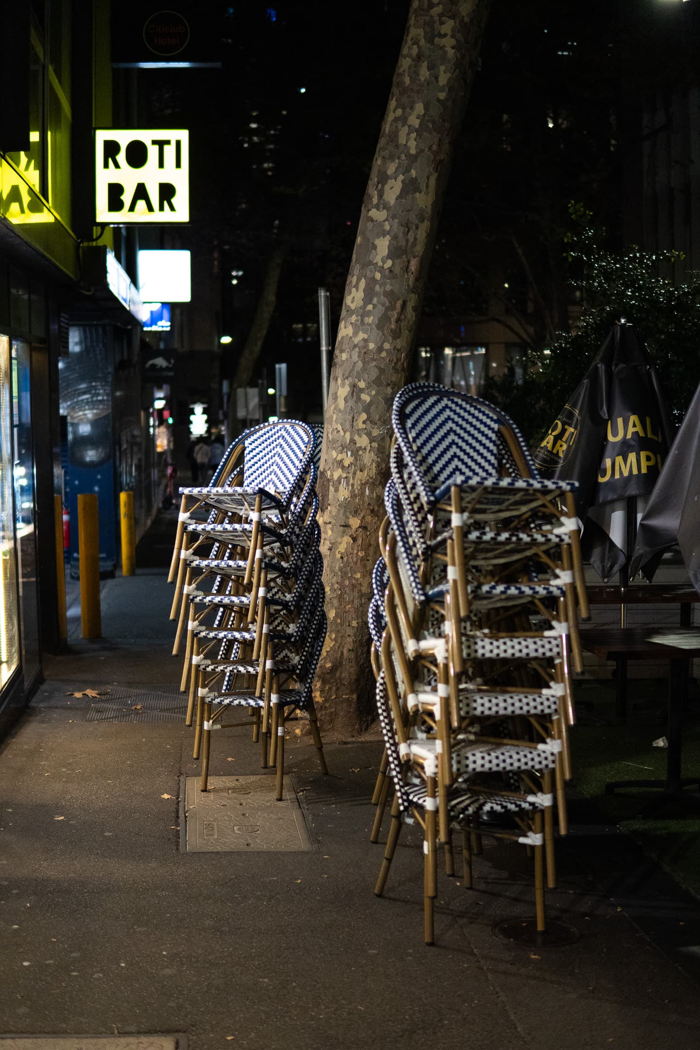 Stacked chairs beside a tree on a city sidewalk at night, with a lit sign for a ROTI BAR in the background