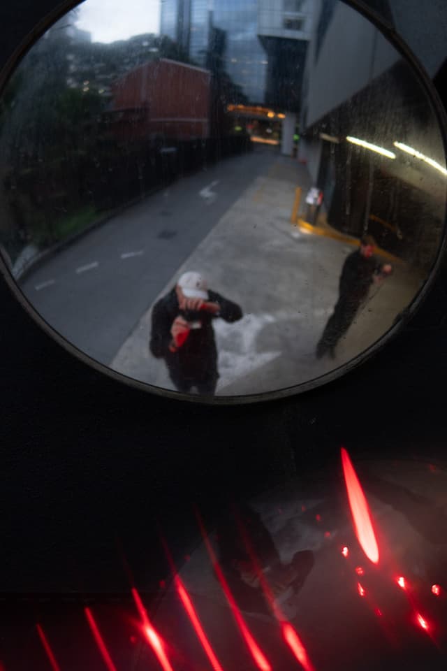 A convex mirror reflects an urban scene with a person taking a photo, another person standing nearby, and a glimpse of buildings and a street, with red light accents at the bottom edge of the mirror