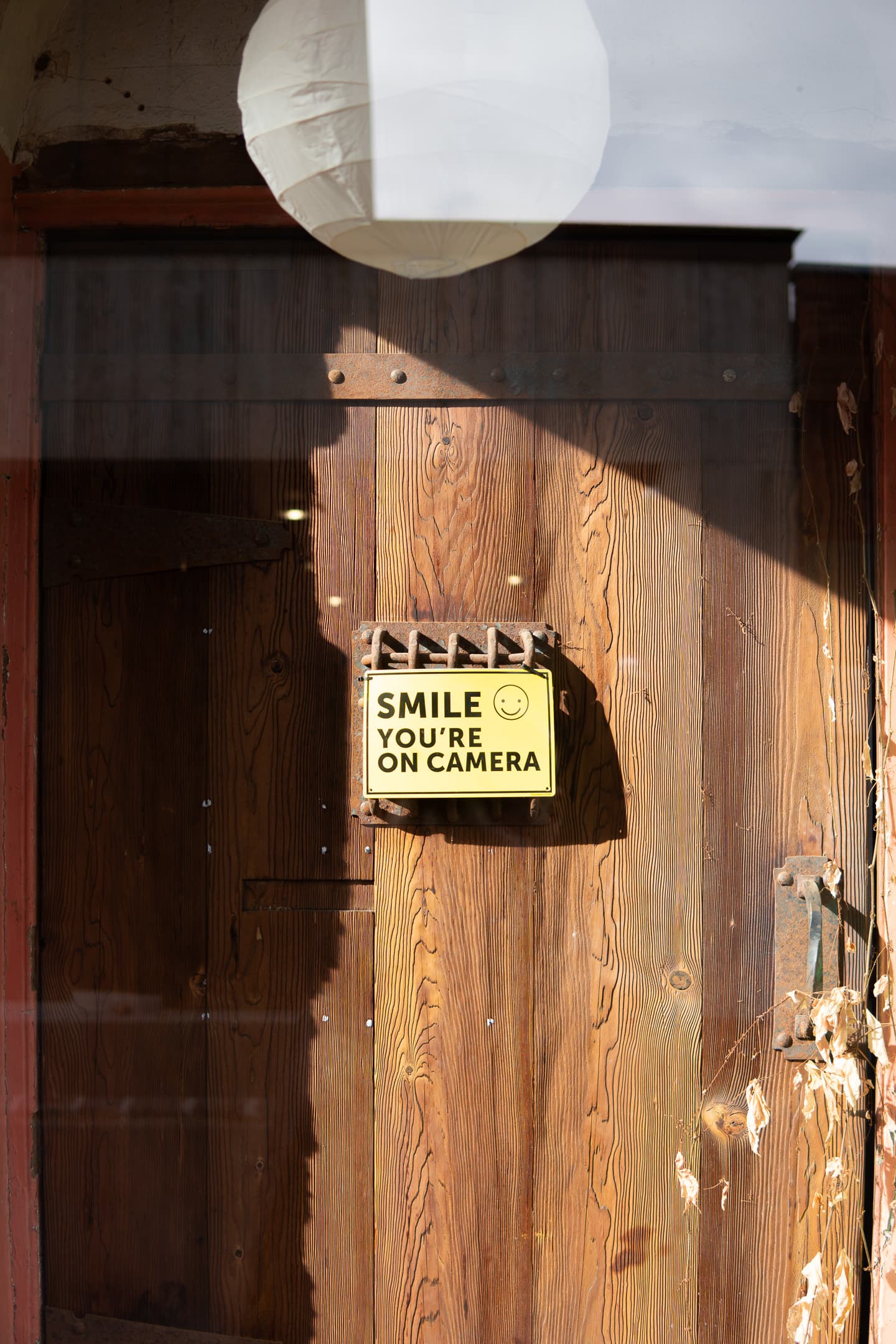 A weathered wooden door with a sign that reads SMILE YOU'RE ON CAMERA featuring a smiley face, with sunlight casting shadows on the surface