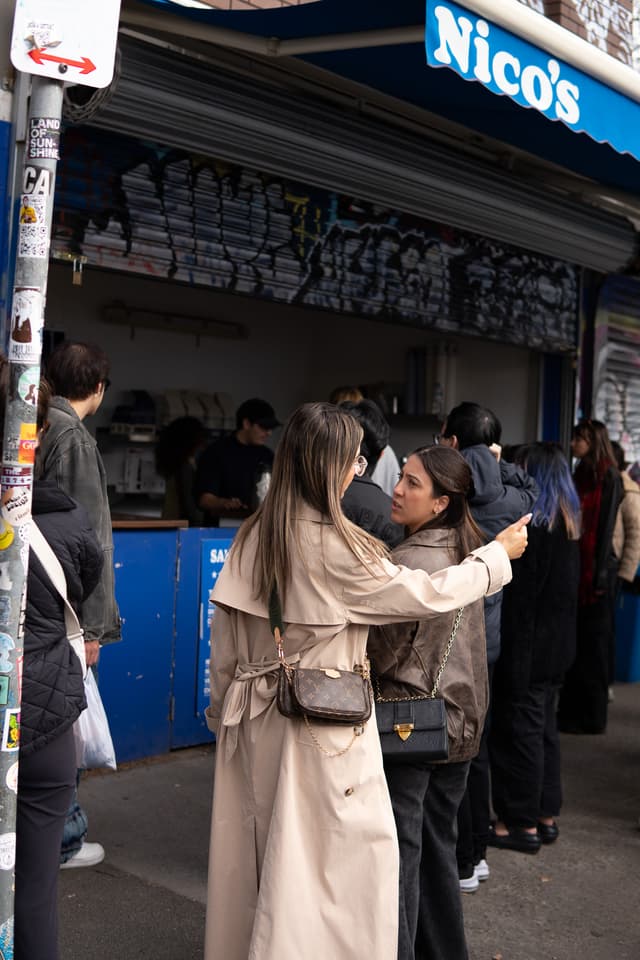 Two individuals are engaged in a conversation in front of a food stand named Nico's, with other people queuing in the background